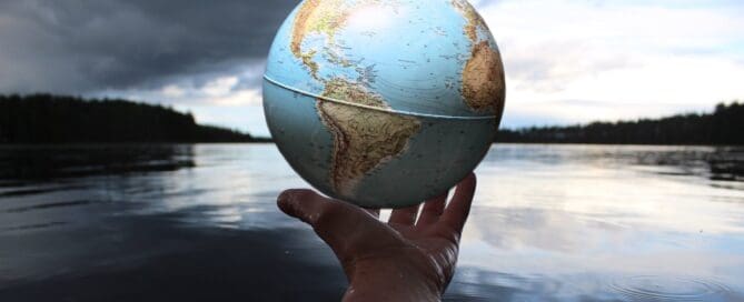 person holding a globe in the palm of their hand with a lake in the background